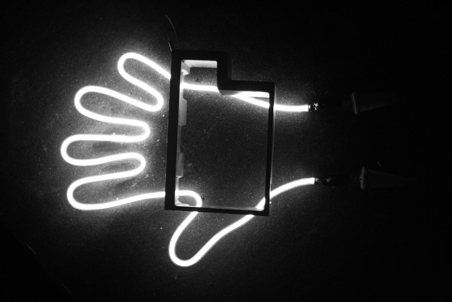 La imagen muestra una instalación luminosa con un diseño moderno y minimalista. Sobre un fondo oscuro, se observa una figura de neón blanco que representa la silueta de una mano abierta, con los dedos extendidos en un trazo continuo y curvado.  En la parte superior derecha de la mano, hay una estructura rectangular tridimensional que parece una especie de marco o contorno de un objeto, interrumpiendo parcialmente el diseño del neón. La combinación de la luz brillante con el fondo oscuro crea un fuerte contraste, destacando la forma de la mano y el objeto.  El efecto de iluminación del neón proyecta un resplandor en la superficie de fondo, lo que añade profundidad y un toque artístico a la composición. La imagen evoca un estilo contemporáneo, posiblemente relacionado con arte, diseño gráfico o señalética moderna.