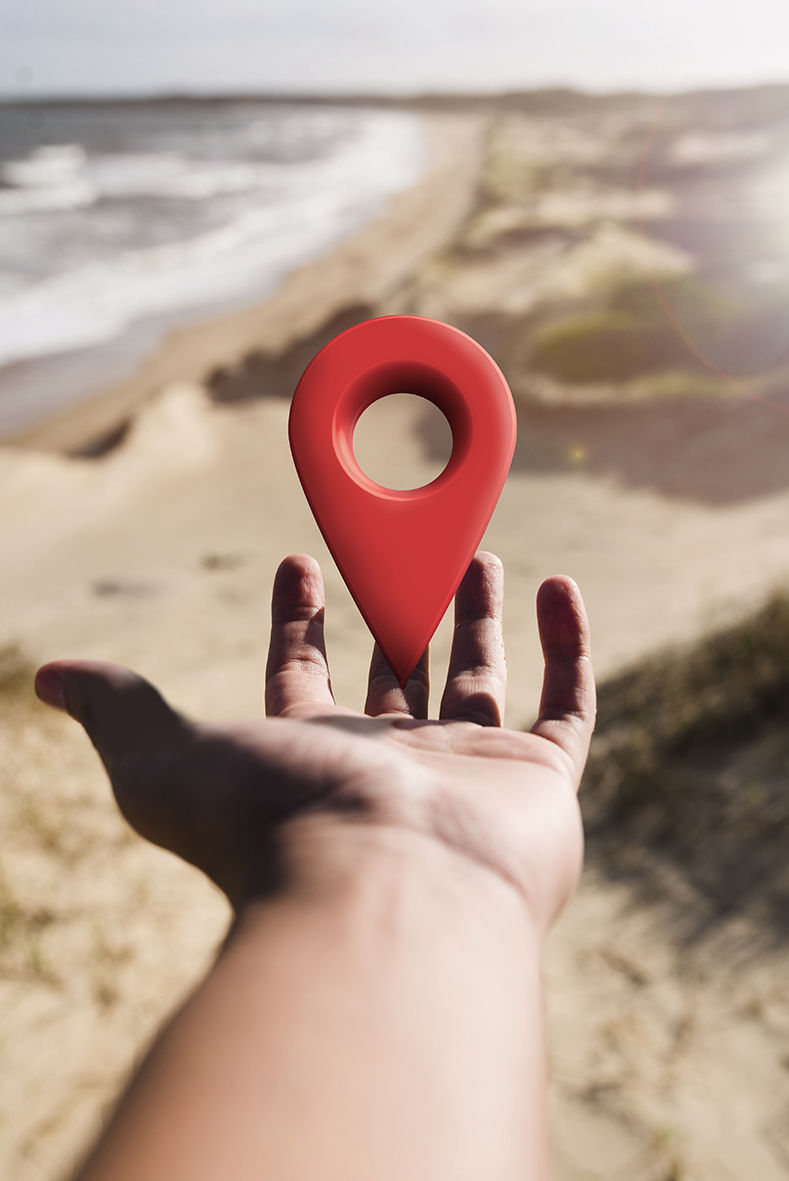 La imagen muestra una mano extendida con la palma hacia arriba, sosteniendo virtualmente un icono de ubicación rojo en 3D (similar al marcador de Google Maps). El fondo es un paisaje natural con tonos cálidos y difuminados, lo que crea una sensación de profundidad y enfoque en el marcador de ubicación. La composición transmite un mensaje de ubicación, geolocalización o selección de un punto estratégico. La iluminación y el desenfoque del fondo resaltan el elemento central de la imagen.